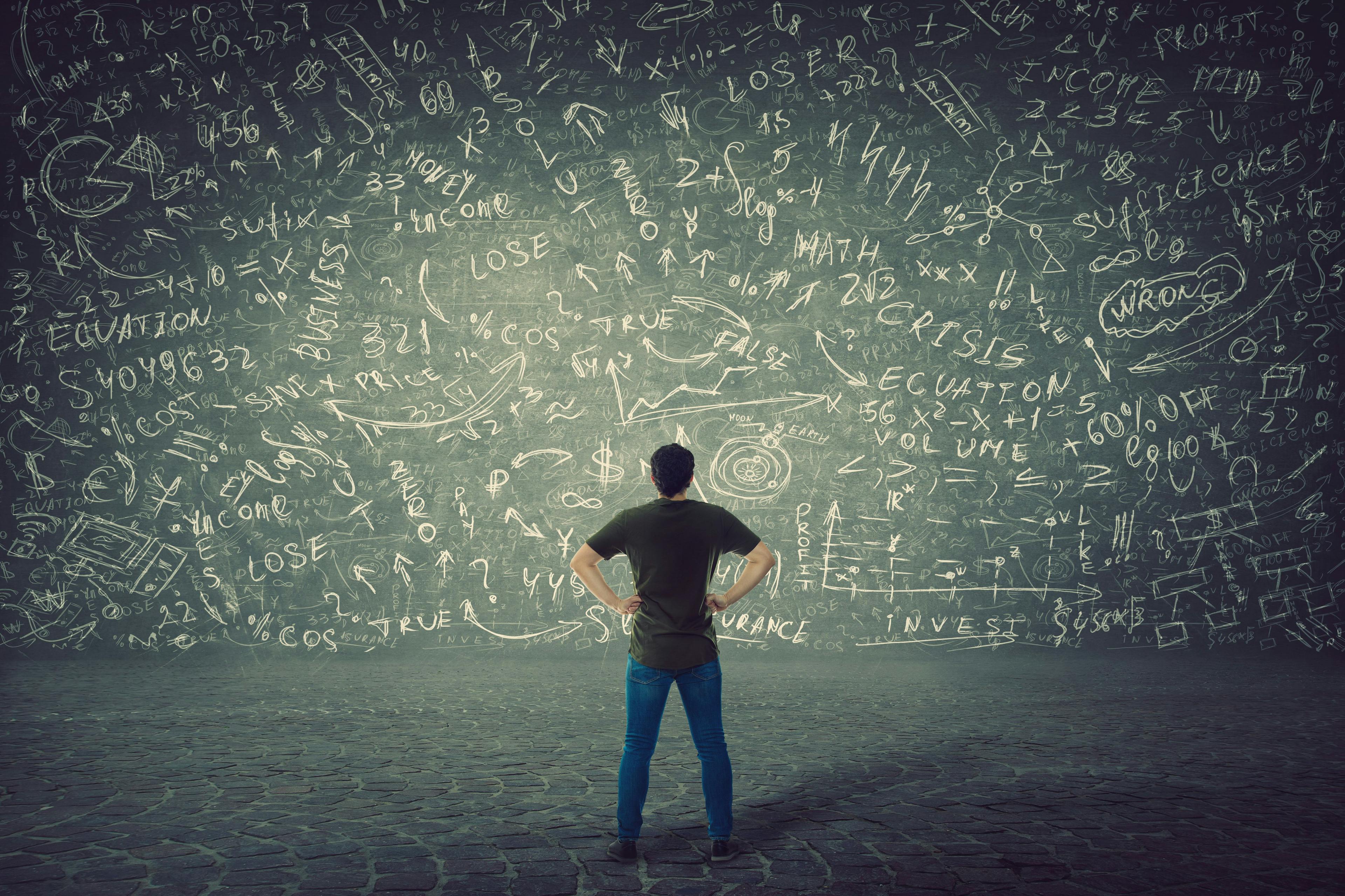 Rear view of puzzled student, hands on hips, stands in front of a huge blackboard try to solve hard mathematics calculation, formula and equations. Project ideas thinking