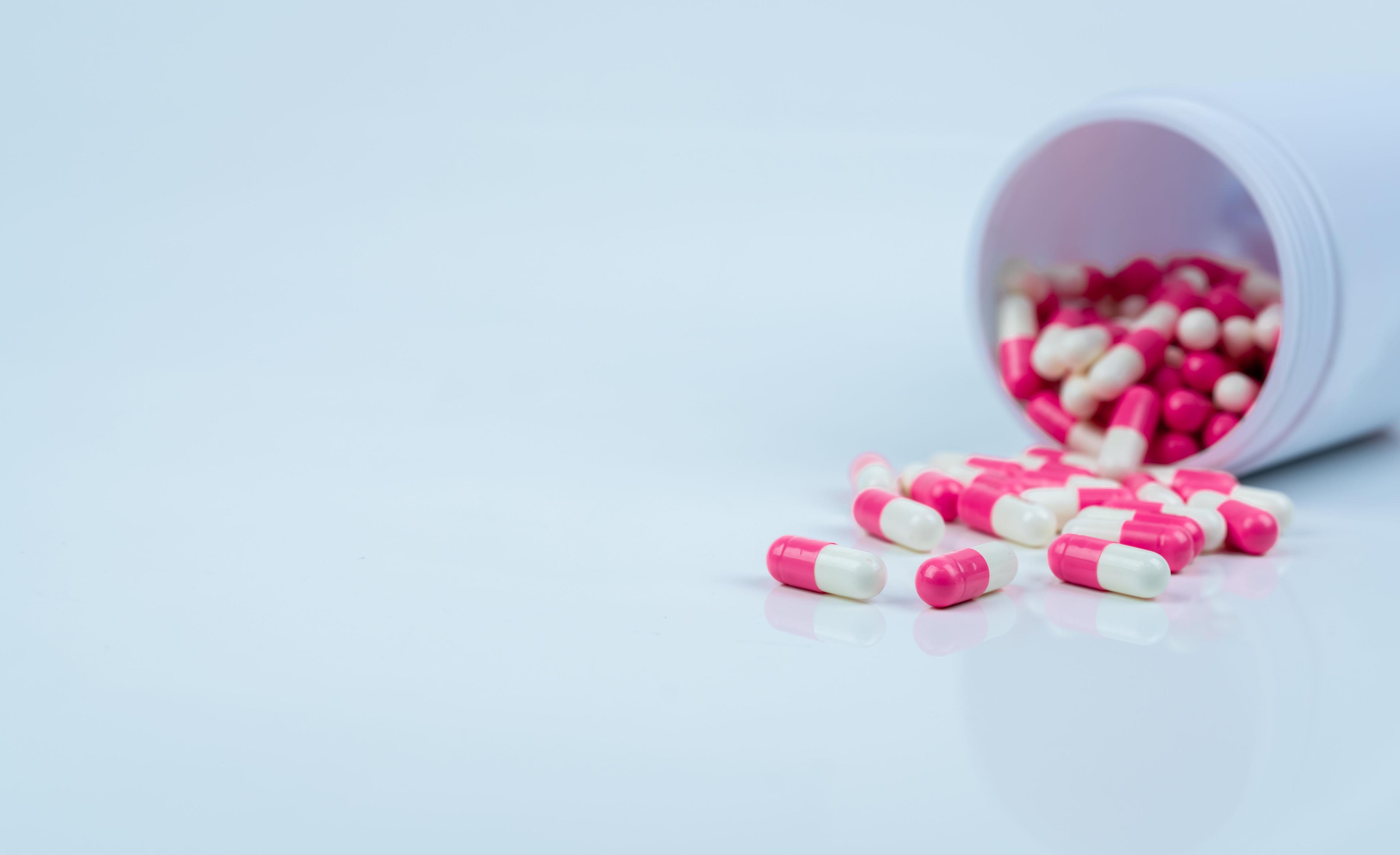 Pink-white capsule pills spread out of drug bottle. Antipsychotic drug. Capsule medicine for treatment depression. Anti-anxiety drug. Global healthcare. Pharmacy background. Pharmaceutical industry. | Image Credit: © Artinun - stock.adobe.com