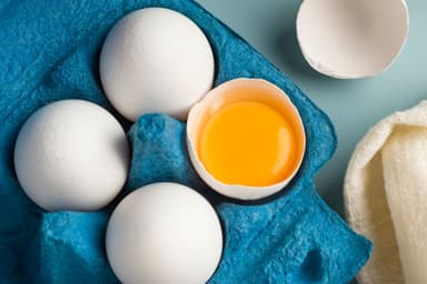 Close-up of chicken fresh eggs in eco-packaging on a blue background. Broken egg with yolk in the shell. Farm natural products. Top view. | Image Credit: © TATIANA